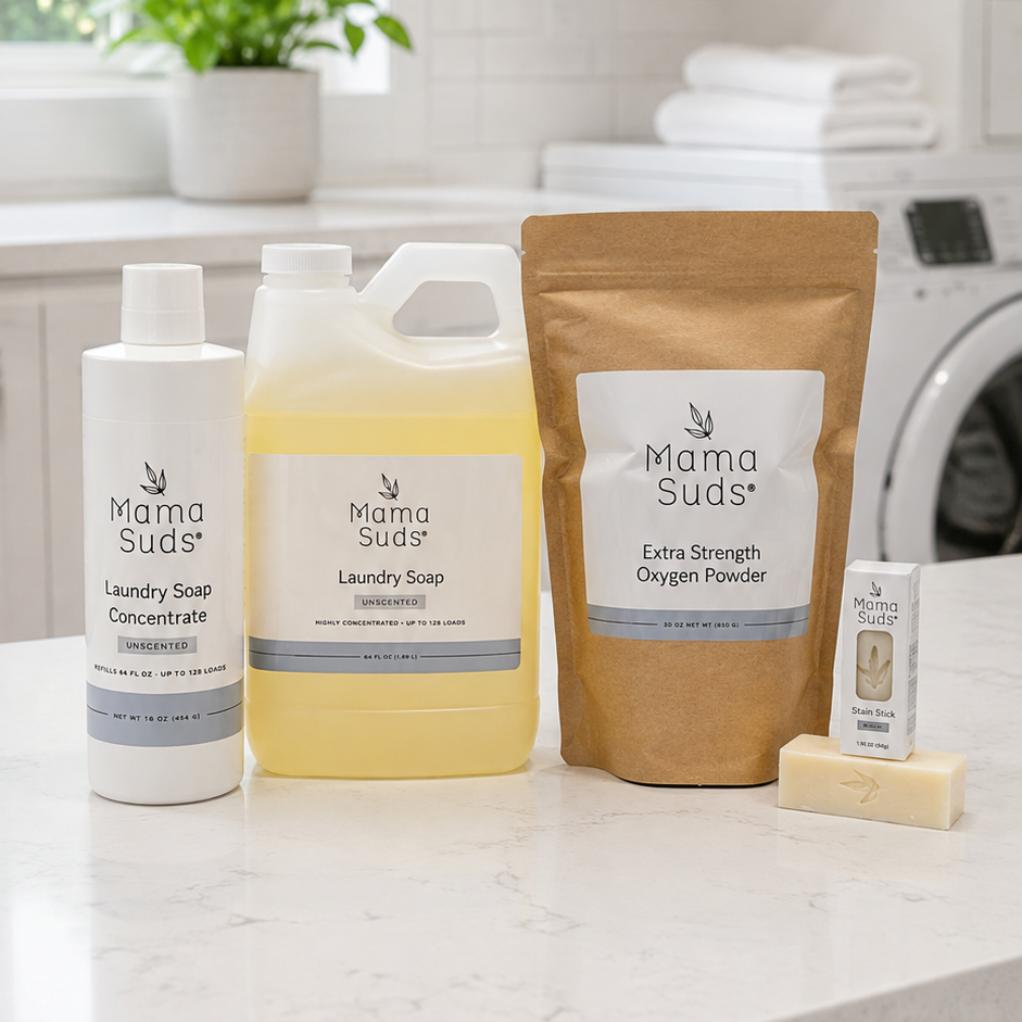 A set of Mama Suds laundry care products arranged on a white marble countertop in a bright, modern laundry room. The products include a bottle of laundry soap concentrate, a large jug of laundry soap, a stand-up pouch of extra strength oxygen powder, and a small bar of soap.