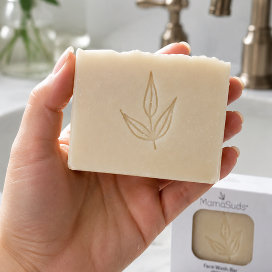 A hand holds a rectangular bar of natural soap with an embossed leaf design. The soap bar is shown in front of a bathroom sink and faucet, with a branded product box and a folded towel visible in the background.