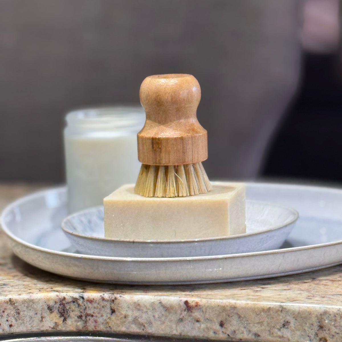 A wooden-handled dish brush with natural bristles resting on a rectangular bar of soap, set on stacked ceramic plates on a granite countertop.