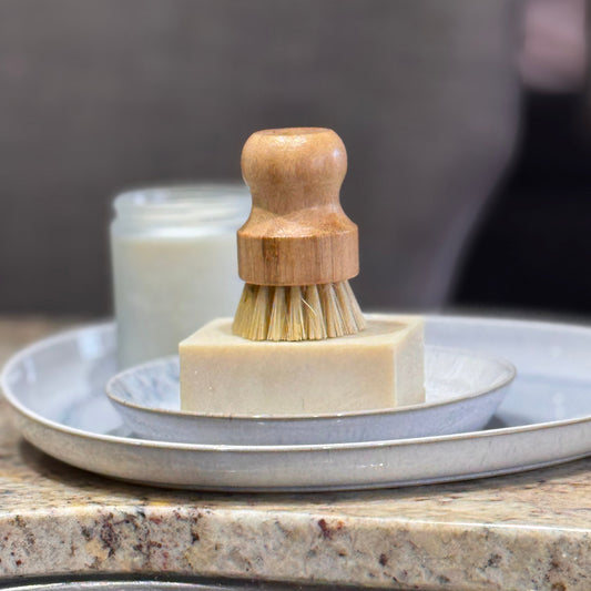 A wooden-handled dish brush with natural bristles resting on a rectangular bar of soap, set on stacked ceramic plates on a granite countertop.