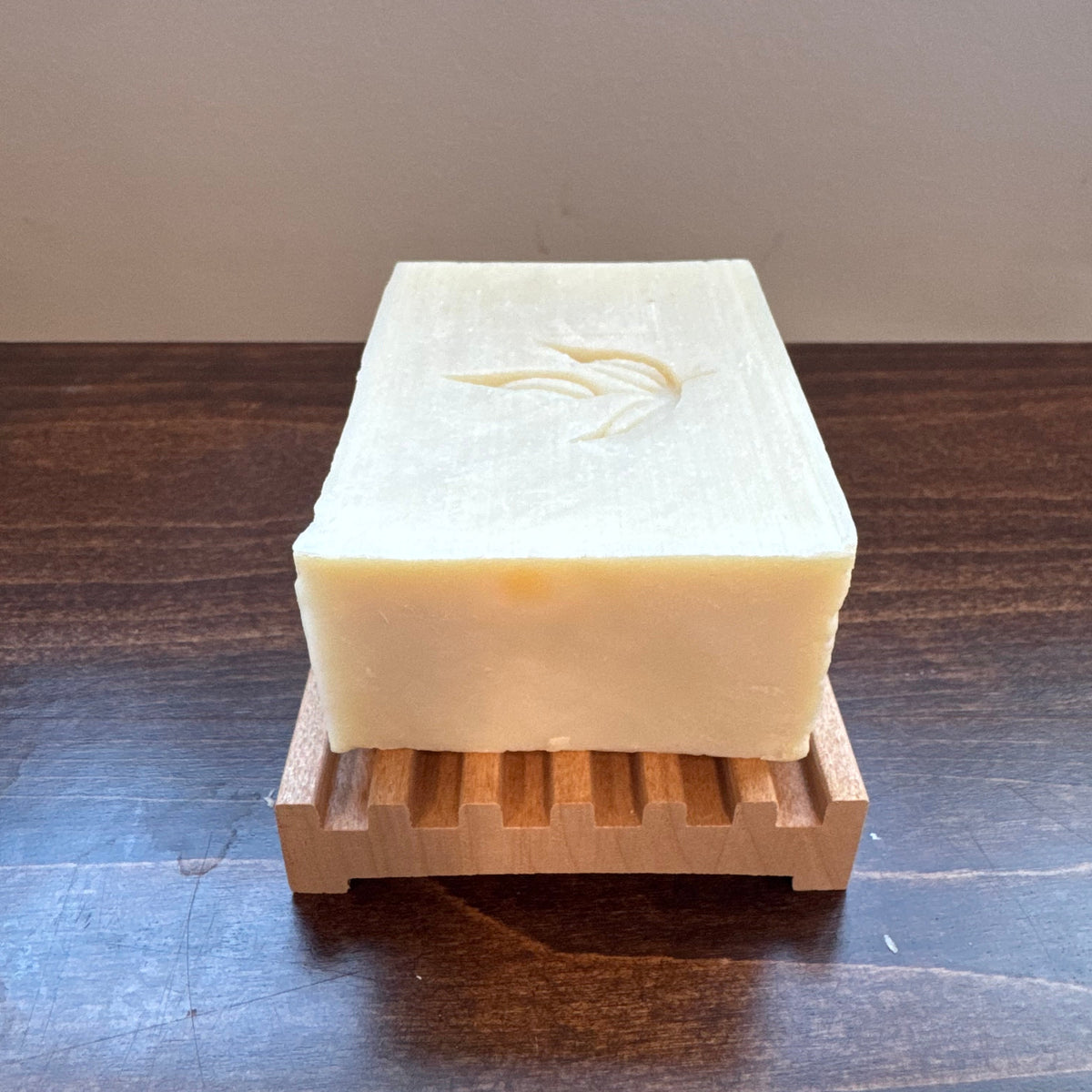 A rectangular bar of creamy off-white soap with a small embossed bird design on top, resting on a small wooden soap dish over a dark wooden surface.