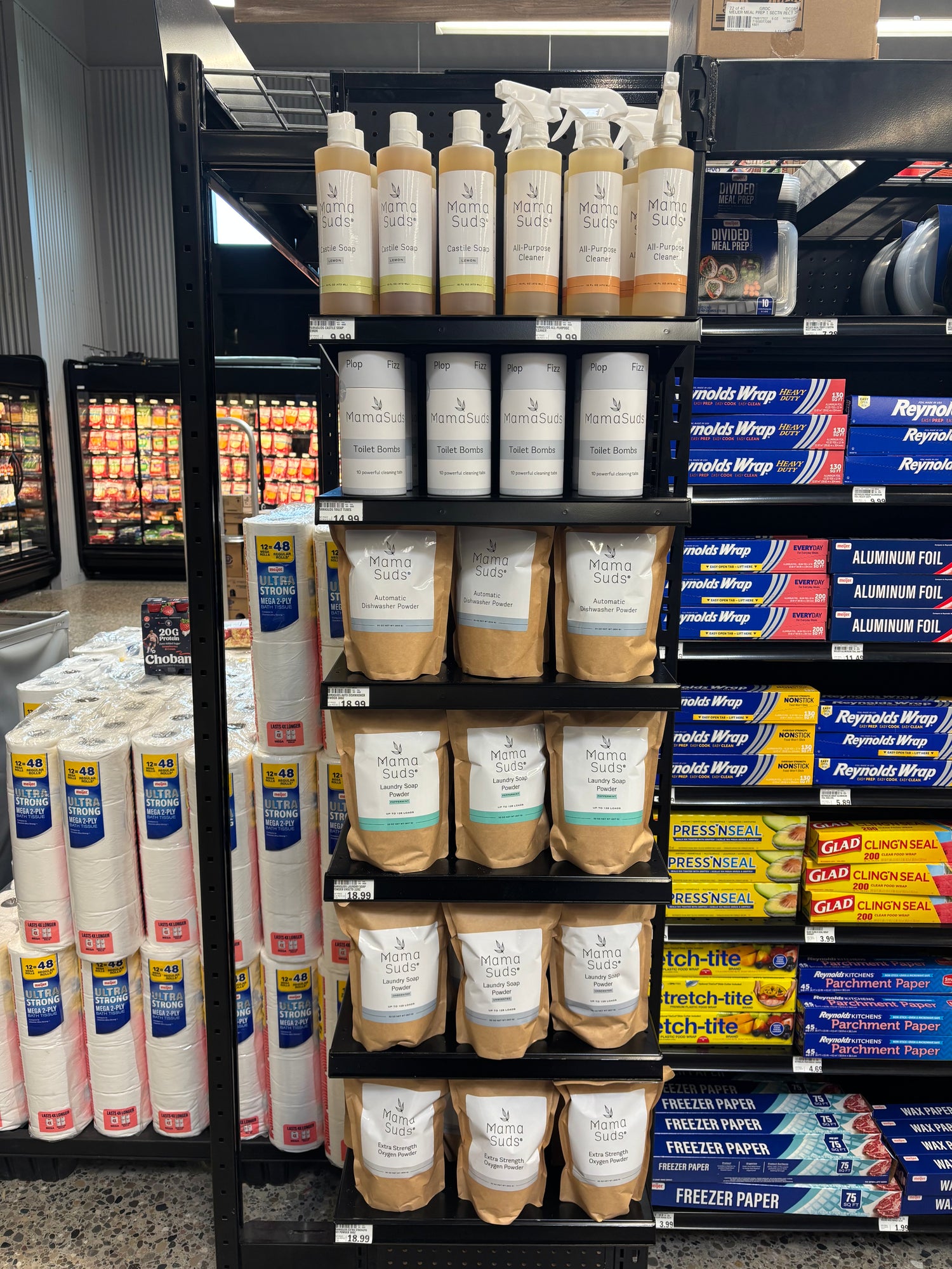 A retail store shelf stocked with various household cleaning products, including bottles of dish soap, spray cleaners, paper towels, and boxes of Reyna's aluminum foil and freezer paper.
