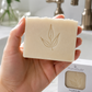 A hand holds a rectangular bar of natural soap with an embossed leaf design. The soap bar is shown in front of a bathroom sink and faucet, with a branded product box and a folded towel visible in the background.