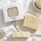 MamaSudis Face Wash Bar shown with its white packaging box, alongside two natural cream-colored soap bars embossed with a leaf design, styled on a white textured fabric with a wooden brush, a bowl of bath sponge, and dried flowers.