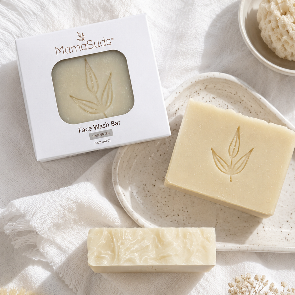 MamaSudis Face Wash Bar shown with its white packaging box, alongside two natural cream-colored soap bars embossed with a leaf design, styled on a white textured fabric with a wooden brush, a bowl of bath sponge, and dried flowers.