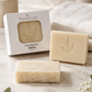 MamaSuds Face Wash Bar shown with its white box packaging and two cream-colored bars, styled on a marble surface with a folded towel, soap dispenser, and small flowers.