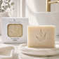 A bar of MamaSuds Face Wash Bar soap sits on a white dish in a bathroom setting, with its white retail box visible behind it on a marble countertop.