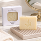 A bar of MamaSuds Face Wash Bar soap in a pale cream color with a gold leaf embossing, displayed on a textured beige soap dish next to its white packaging box. The scene is styled with a folded cream towel, a round gold-framed mirror, and soft white flowers on a light marble surface.