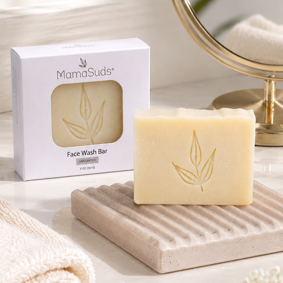 A bar of MamaSuds Face Wash Bar soap in a pale cream color with a gold leaf embossing, displayed on a textured beige soap dish next to its white packaging box. The scene is styled with a folded cream towel, a round gold-framed mirror, and soft white flowers on a light marble surface.