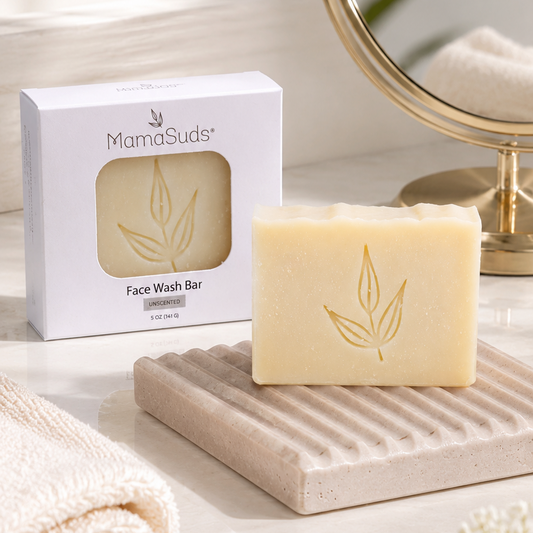 A bar of MamaSuds Face Wash Bar soap in a pale cream color with a gold leaf embossing, displayed on a textured beige soap dish next to its white packaging box. The scene is styled with a folded cream towel, a round gold-framed mirror, and soft white flowers on a light marble surface.